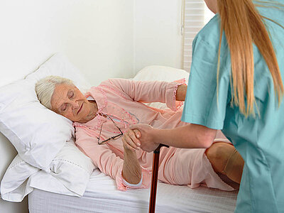 Auf dem Bild sieht man eine Person in türkiser Kleidung, die einer anderen Person auf einem Bett hilft und deren Fuß hält. Es veranschaulicht die Bedeutung von Fürsorge und Unterstützung im Gesundheitswesen.