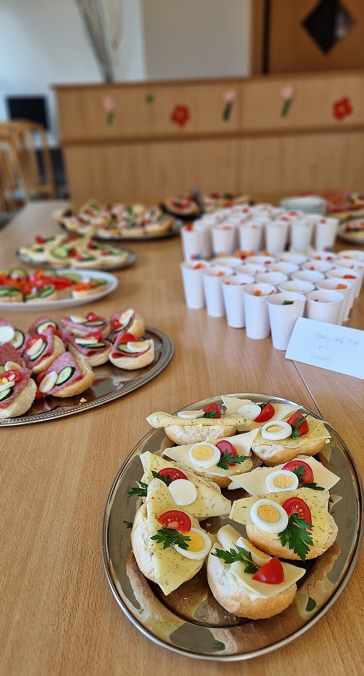Berufspädagogische Pflichtfortbildung an der Euro Akademie Pößneck Belegete Weißbrotbrötchen liegen auf einem silbernen Tablett und sind belegt mit Käse, Tomate, Gurke und verschiedener Wurst. Dort herum stehen gestapelte Becher.