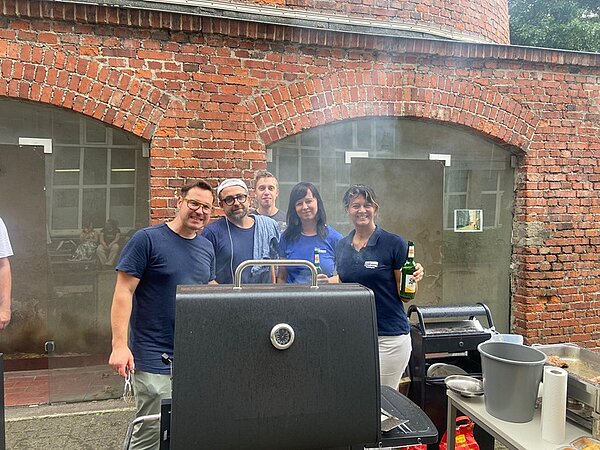 Sommerliches Abschlussfest an der Euro Akademie Jena Lehrkräfte stehen hinter einem großen Grill, es raucht, alle lächeln gut gelaunt für ein Foto in die Kamera.