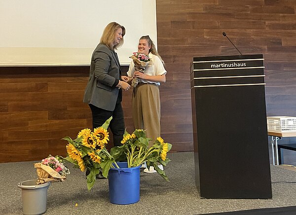 Eine Frau überreicht einer anderen Frau einen Blumenstrauß auf einer Bühne. Beide lächeln. Im Hintergrund ist ein Pult mit der Aufschrift "martinushaus" zu sehen. Auf dem Podium stehen weitere Blumensträuße mit Sonnenblumen.
