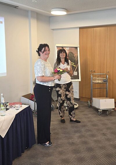 Zwei Frauen stehen lächelnd nebeneinander in einem Konferenzraum. Die Frau links trägt ein weißes, gemustertes Oberteil und eine schwarze Hose und hält einen Blumenstrauß in den Händen, während die Frau rechts ein weißes Oberteil und eine schwarz-beige gemusterte Hose trägt.