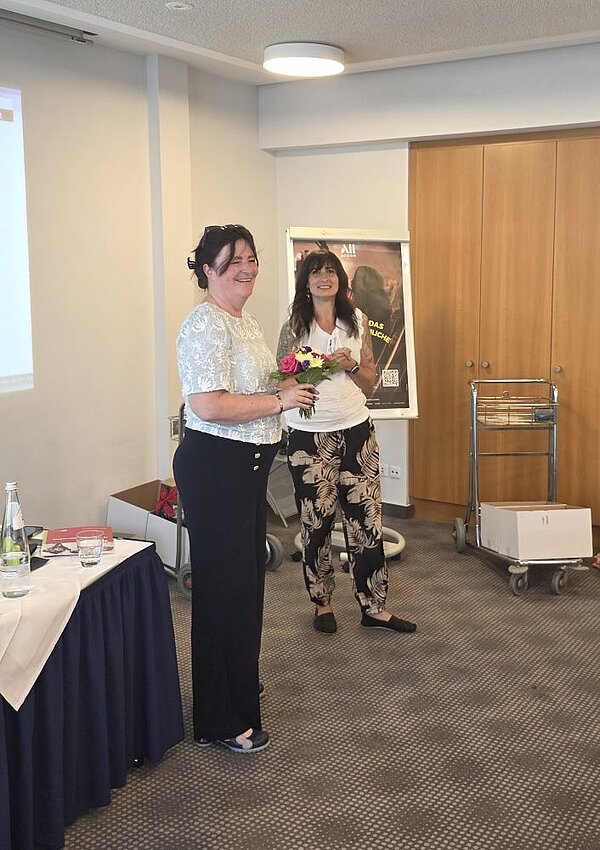 Zwei Frauen stehen lächelnd nebeneinander in einem Konferenzraum. Die Frau links trägt ein weißes, gemustertes Oberteil und eine schwarze Hose und hält einen Blumenstrauß in den Händen, während die Frau rechts ein weißes Oberteil und eine schwarz-beige gemusterte Hose trägt.