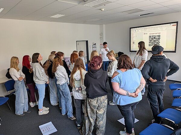 Die Auszubildenden stehen im Klassenraum vor einem großen Bildschirm. Auf dem Bildschirm sind ein QR-Code und eine Präsentationsfolie zum Filmprojekt zu sehen.