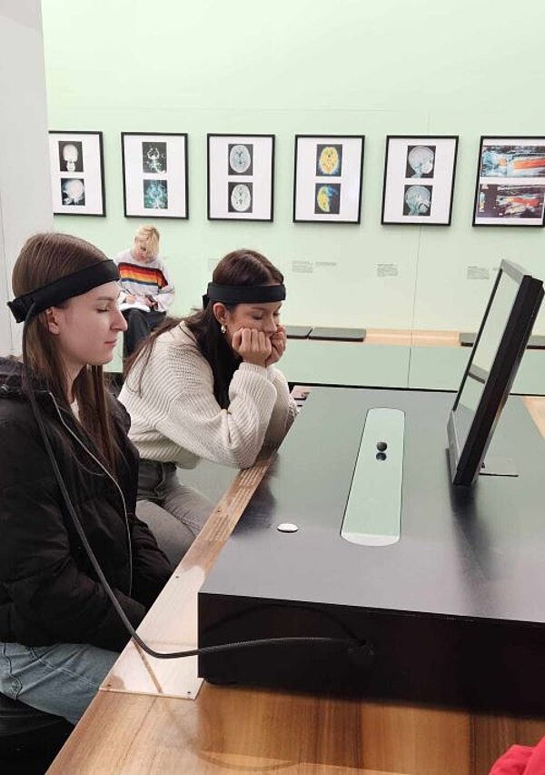 Bewegung und Balance im Hygienemuseum Zwei junge Frauen sitzen an einem Tisch auf dem ein großes schwarzes Gerät steht, auf dem ein Monitor angebracht ist. Die beiden Frauen sind mit einem dicken Kabel, das an ihren Köpfen befestigt sind mit dem Gerät verbunden. Sie haben die Augen geschlossen.