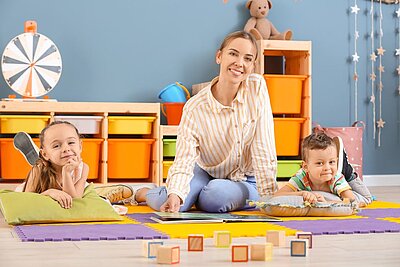 Eine Erzieherin sitzt mit zwei fröhlichen Kindern auf dem Boden eines bunten Kindergartenraums. Sie lächelt in die Kamera, während die Kinder entspannt auf Kissen liegen. Im Hintergrund befinden sich Regale mit Spielzeug, Bauklötzen und ein Kuscheltier, die eine warme und einladende Atmosphäre schaffen.