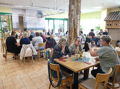 Gut besuchter Gastraum mit mehreren Gruppen an Holztischen, die gemeinsam essen, trinken und sich unterhalten.