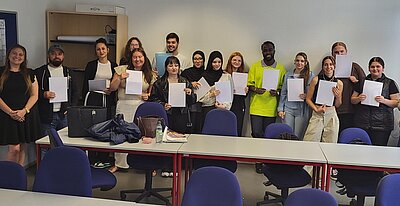 Gruppenfoto von jungen Erwachsenen in einem Klassenraum, die ihre Zeugnisse oder Teilnahmebescheinigungen präsentieren. Die Teilnehmenden stehen nebeneinander und lächeln. Auf den Tischen vor ihnen liegen Taschen, Getränke und Unterlagen.