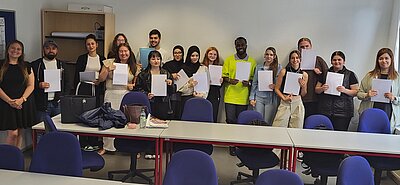 Gruppenfoto von jungen Erwachsenen in einem Klassenraum, die ihre Zeugnisse oder Teilnahmebescheinigungen präsentieren. Die Teilnehmenden stehen nebeneinander und lächeln. Auf den Tischen vor ihnen liegen Taschen, Getränke und Unterlagen.