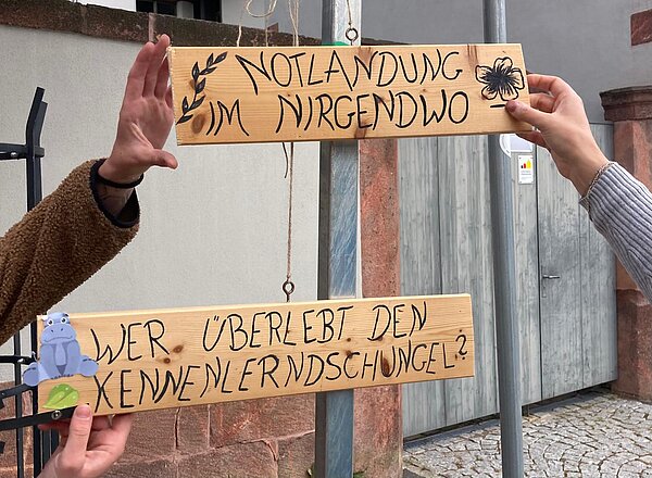 Kennenlerntag der Euro Akademie Rochlitz Zwei Holzschilder mit der Aufschrift „Notlandung im Nirgendwo“ und „Wer überlebt den Kennenlerndschungel?“. Sie sind handbemalt und werden von zwei Personen gehalten.