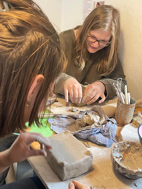 Zwei junge Frauen arbeiten konzentriert an ihren Tonobjekten. Eine formt eine rechteckige Schale, während auf dem Tisch Modellierwerkzeuge, Ton und eine Schüssel mit Tonschlicker liegen.