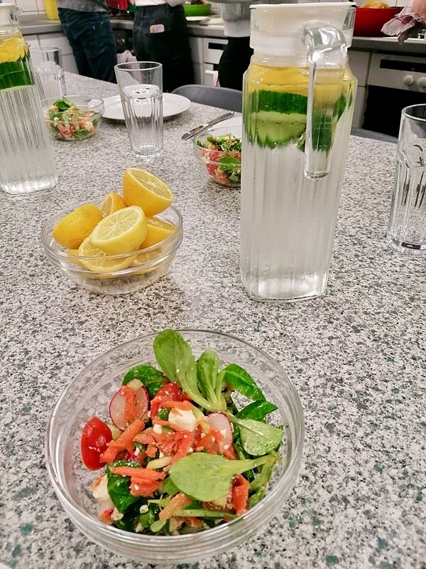 An die Töpfe, fertig, los! Salat in Glasschüssel steht, gemeinsam mit halbierten Zitronen und einer Karaffe mit Limonen- und Zitrionenscheiben, auf dem Tisch.