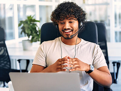 Ein lächelnder Mann mit lockigem Haar und Headset sitzt an einem Schreibtisch in einem Büro. Er trägt ein helles T-Shirt und hat die Hände vor sich gefaltet. Vor ihm steht ein Laptop, und im Hintergrund sind weitere Büromöbel und eine Pflanze zu sehen.