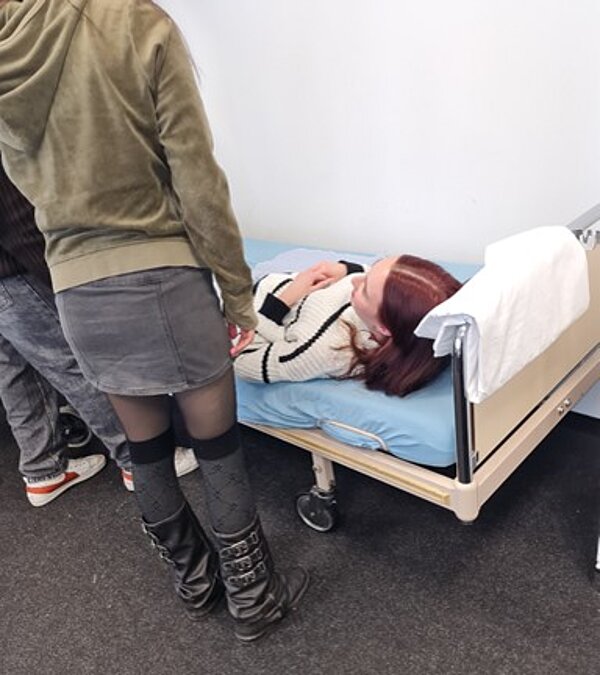 Pflege trifft Soziales Eine Person mit roten Haaren liegt auf einem Pflegebett mit blauer Matratze und hält eine Hand auf der Brust. Sie trägt einen weißen Pullover mit schwarzen Streifen. Neben dem Bett steht eine weitere Person in einem grünen Hoodie, einem kurzen Rock und schwarzen Stiefeln. Im Hintergrund sind weitere Personen mit Sneakers zu sehen, die sich an der Pflegesituation beteiligen.