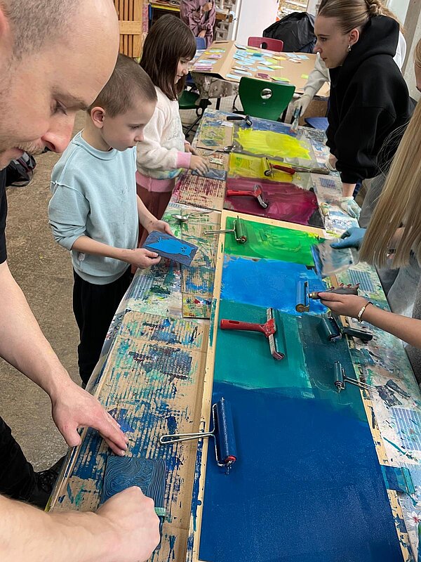 Lange Druckstraße: Kinder rollen gelbe, grüne und blaue Farbe mit Walzen auf Druckplatten auf; Betreuung durch Erwachsene.