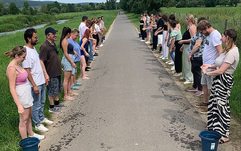 Eine Gruppe von Menschen steht in zwei Reihen entlang eines schmalen, ländlichen Weges. Sie halten sich an den Händen und bilden eine Art menschliche Kette. Jede Person hat einen Eimer bei sich. Der Weg ist von grünen Feldern und Bäumen umgeben, und der Himmel ist klar. Die Szene wirkt wie eine Gemeinschaftsaktion oder ein Event im Freien.