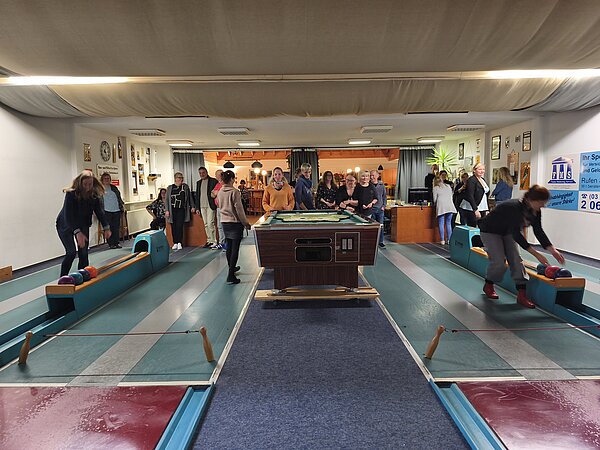 Zwei aktive Kegelbahnen: Teilnehmende werfen gleichzeitig Kugeln, andere beobachten dahinter; lebhafte Bewegung und gesellige Stimmung.