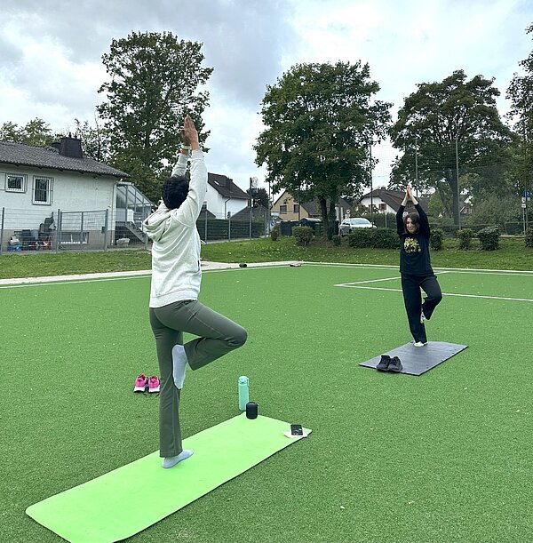 Together in motion – Bewegung, Spaß und Teamgeist Zwei Personen machen Yoga auf einem grünen Kunstrasenplatz im Freien. Beide stehen auf einer Yoga-Matte und führen eine Übung im Stehen mit einem Arm nach oben gestreckt aus. Im Hintergrund sind Bäume und ein Wohnhaus zu sehen.