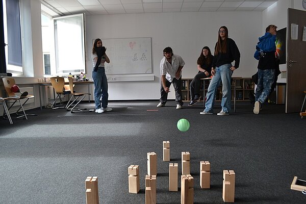 Besucherinnen und Besucher des Tages der offenen Tür an der Euro Akademie Würzburg stehen in einem Klassenraum, in dem die Stühle und Tische beiseite geschoben wurden, und spielen Wikingerschach. 