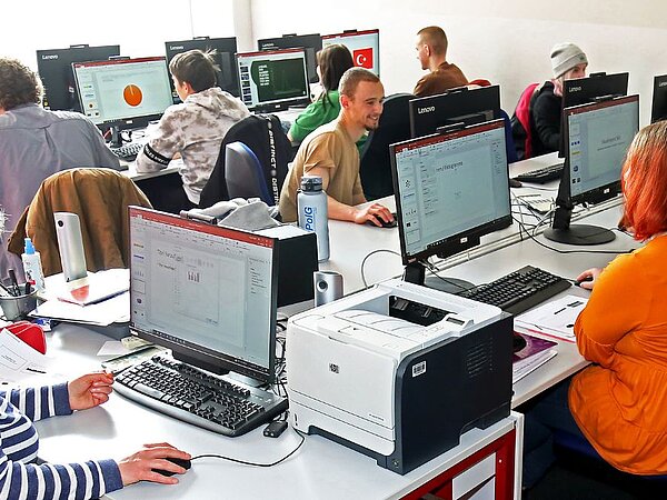 Eine Gruppe von Menschen verschiedenen Alters sitzt in einem Computerraum und arbeitet konzentriert an PCs. Die Personen schauen auf die Bildschirme. Der Raum ist hell und modern eingerichtet, mit mehreren Reihen von Tischen und Computern."