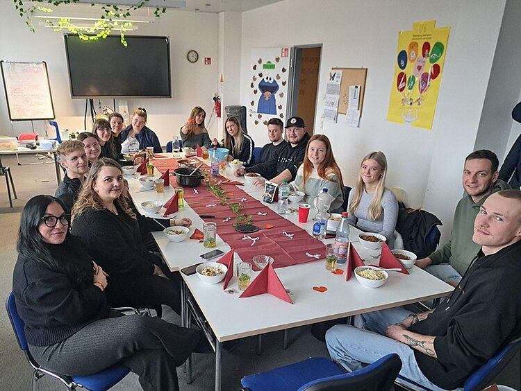 Halbjahresinformation Schülerinnen und Schüler sitzen mit ihrer Lehrerin gut gelaunt an einem langen gedeckten Frühstückstisch.