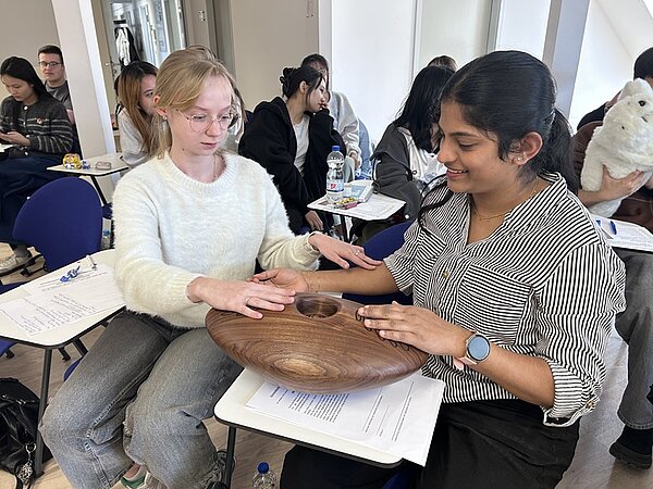 Zwei Auszubildende sitzen sich in einem Klassenraum gegenüber. Beide haben die Hände auf einer runden, flachen Holzscheibe mit einer Vertiefung in der Mitte abgelegt. Die eine Person trägt einen hellen Pullover, die andere eine gestreifte Bluse und ein blaues Armband. Auf den Tischen liegen Blätter mit Text und Stiften. Im Hintergrund sitzen weitere Personen auf Stühlen, einige halten Gegenstände in den Händen, darunter ein weißes Plüschtier. Die Szene wirkt ruhig und konzentriert.