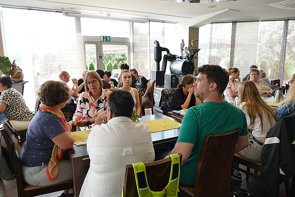 Zahlreiche Ausflugsgäste sitzen in einem hellen Café beziehungsweise Restaurant an mehreren Tischen und unterhalten sich miteinander.