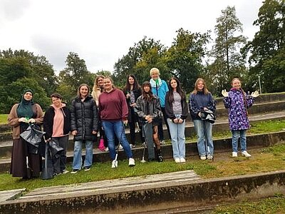 Eine Gruppe von Frauen unterschiedlichen Alters hat sich für das Foto in zwei Reihen aufgestellt. Sie halten Müllsäcke und Müllsammelzangen in den Händen und stehen auf einer begrünten Stufe. Im Hintergrund sind Bäume zu sehen.
