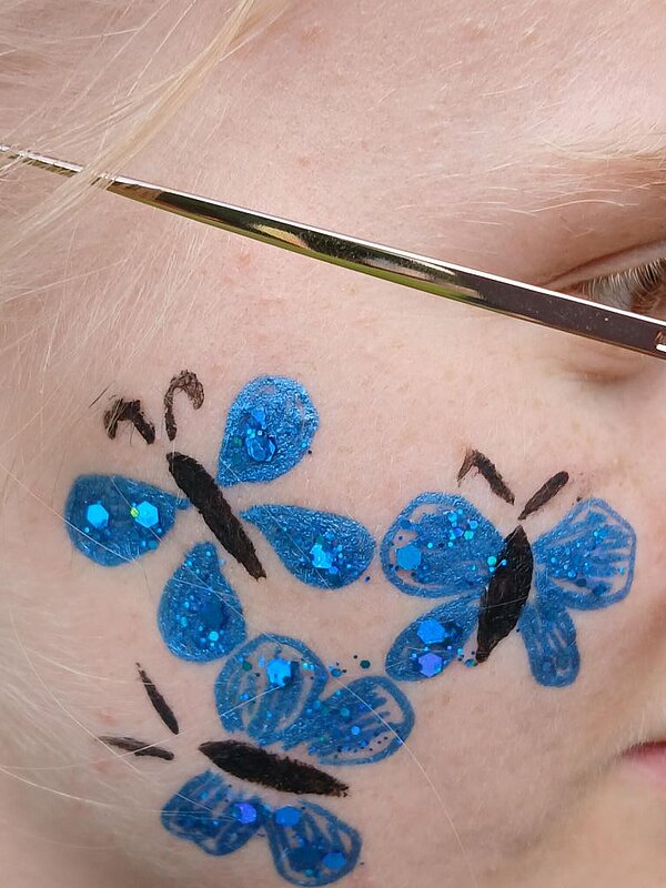  Ein temporäres Tattoo in Form von blauen Schmetterlingen mit schwarzen Körpern und Glitzerakzenten auf dem Gesicht einer Person.