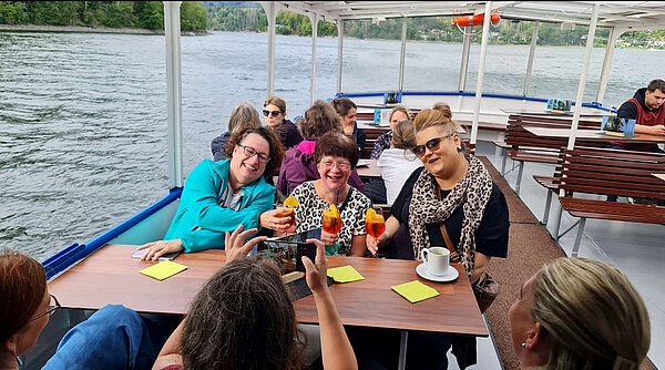 Drei Frauen sitzen während einer Schifffahrt an einem Tisch auf dem Außendeck, lachen und stoßen mit orangefarbenen Getränken an.