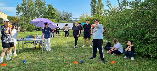 Wikinger-Sportfest: Pflegeazubis erobern die nordische Arena Eine Gruppe von Jugendlichen und jungen Erwachsenen steht auf einer Wiese. Ein junger Mann vorne wirft einen kleinen Ball, während andere um einen Tisch mit lila Sonnenschirm herumstehen.