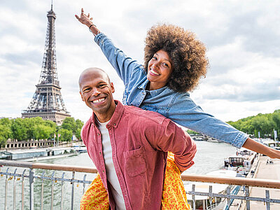 Zwei Personen stehen vor dem Eiffelturm in Paris. Eine hebt die Arme. Die Seine fließt unter ihnen. Helle Wolken am Himmel.