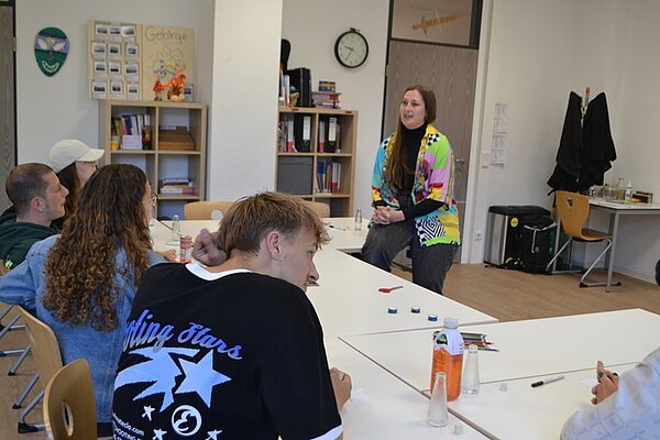 Besucherinnen und Besucher des Tages der offenen Tür an der Euro Akademie Würzburg sitzen in einem Raum und hören der Lehrkraft, die lässig an einen Tisch gelehnt ist und einen farbenfrohen Cardigan trägt, zu. 