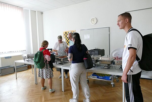 In einem hellen Seminarraum mit großen Fenstern stehen mehrere Personen an einem Buffet. Eine Person in einem gepunkteten Rock mit grünem Rucksack wird von zwei Personen hinter den Tischen bedient. Zwei weitere Personen tragen Rucksäcke oder Taschen und warten in der Schlange.