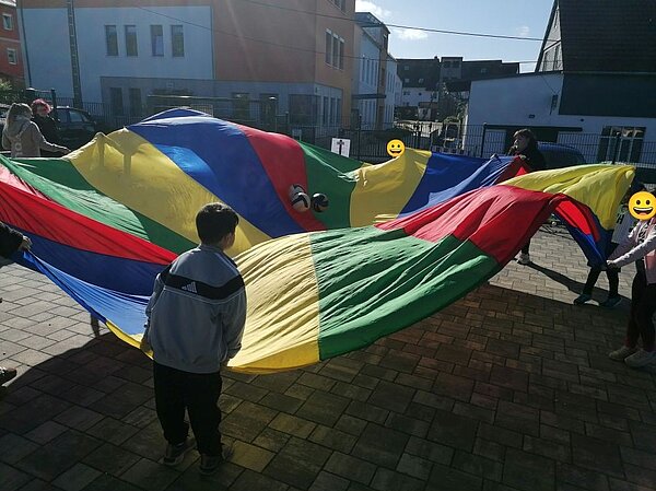 Kinder und Erwachsene halten gemeinsam einen großen Fallschirm in Gelb, Grün, Rot und Blau hoch; fröhliche Stimmung auf einem sonnigen Platz vor bunten Gebäuden.