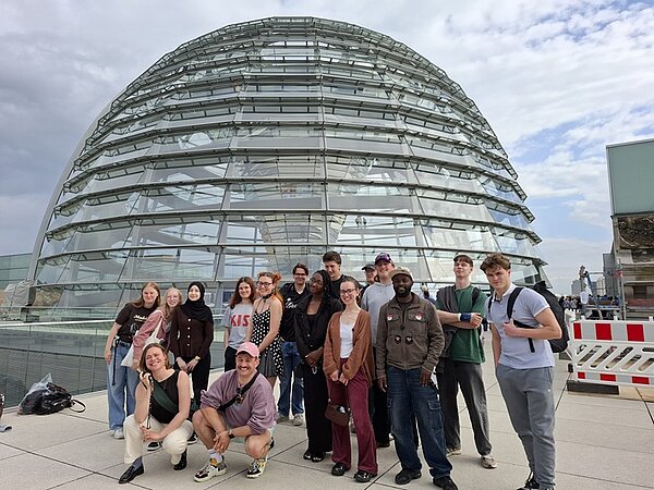 Die Azubi-Gruppe steht auf einer Dachterrasse vor einer großen, gläsernen Reichstagskppel mit geschwungenen Metallstreben und posieren für ein Erinnerungsfoto. Alle sind locker nebeneinander aufgestellt, einige stehen, andere hocken. Im Hintergrund ist der Himmel bewölkt. Rechts im Bild befindet sich eine rot-weiße Absperrung.