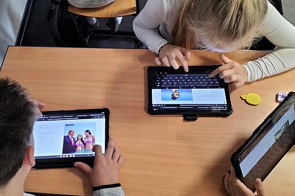 Drei Schülerinnen und Schüler sitzen an einem Holztisch und arbeiten mit Tablet-Computern. Die Bildschirme zeigen verschiedene Websites, darunter einen Artikel mit einem Foto von zwei Erwachsenen und einem Kind sowie eine Seite mit einer Tastaturansicht. Die Jugendlichen tragen helle Kleidung und konzentrieren sich auf die Bildschirme. Das Bild entstand im Rahmen eines Medienkompetenz-Projektes der Euro Akademie Görlitz zum Thema Fake News und verdeutlicht den praktischen Umgang mit digitalen Medien im Unterricht.