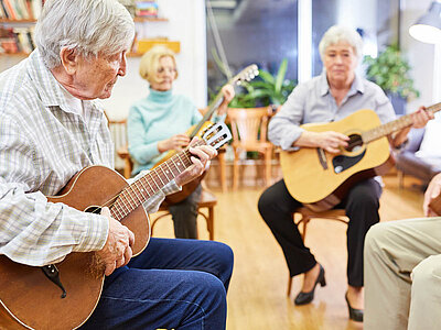 Drei ältere Personen sitzen in einem Raum und spielen Gitarre. Sie wirken konzentriert und genießen das gemeinsame Musizieren.