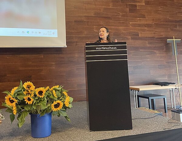 Eine Frau steht an einem Pult mit der Aufschrift "martinushaus" und hält eine Rede. Auf dem Podium stehen Blumenarrangements mit Sonnenblumen.