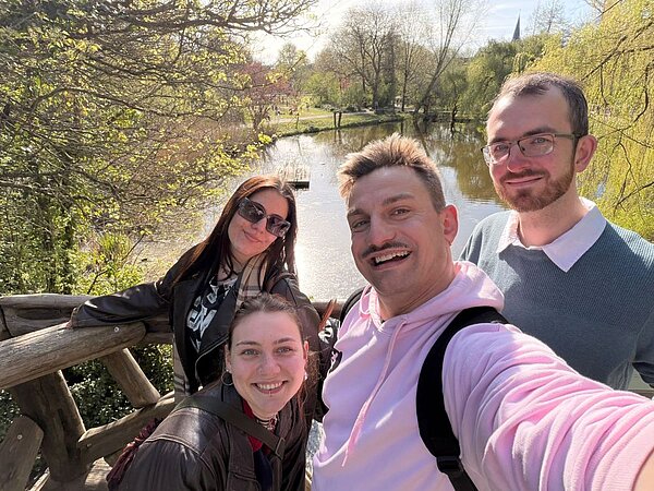 Eine Gruppe von vier lächelnden Personen macht ein Selfie an einem See in einem Park. Die Umgebung ist grün, und im Hintergrund sind Bäume und ein Steg zu sehen.