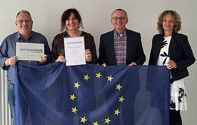 Mitarbeitende und die Leitung der Euro Akademie Hannover posieren, hinter einen großen Europaflagge und mit Urkunde in der Hand, für ein Gruppenfoto zum Re-zertifizierten Titel der Europaschule.