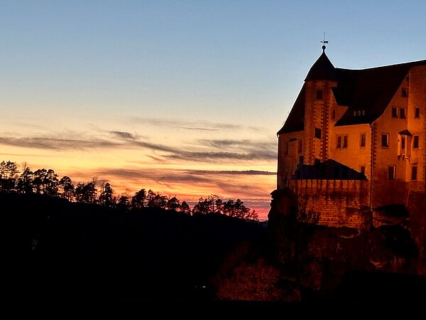 Historisches Schloss auf einem Felsen bei Sonnenuntergang, goldene Lichtstimmung am Himmel mit silhouettiertem Wald im Hintergrund.