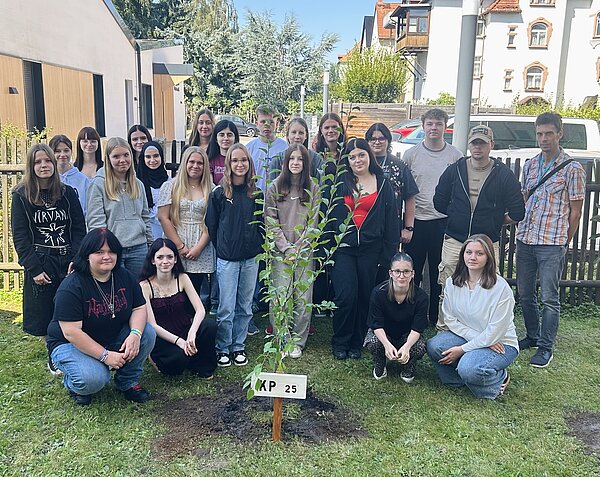 Grüner Schulstart 2025/26 an der Euro Akademie Altenburg Eine Gruppe von Menschen steht im Freien um einen kleinen Baum. Sie posieren lächelnd für ein Gruppenfoto. Der Baum hat ein Schild mit der Aufschrift "KP 25". Die Gruppe besteht aus jungen Erwachsenen und sie stehen auf einem Rasen mit einem Gebäude im Hintergrund.