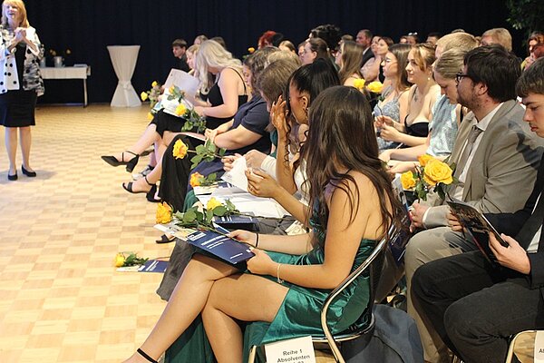 Absolventinnen und Absolventen sitzen im großen Saal auf Stühlen zu ihrer feierlichen Zeugnisübergabe und halten eine gelbe Rose sowie eine Euro Akademie-Broschüre auf dem Schoß und in Händen.