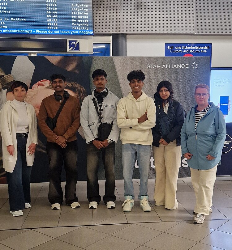 Eine Gruppe von sechs Personen steht in einer Reihe in einem Flughafen. Im Hintergrund sind ein digitales Abflug- und Ankunftsdisplay mit Fluginformationen sowie ein Star Alliance-Schalter und ein Hinweis auf den Zoll- und Sicherheitsbereich zu sehen. Die Personen tragen lockere Kleidung, einige lächeln oder posieren für das Foto.