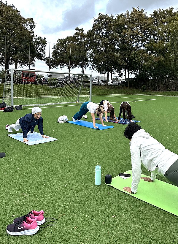 Together in motion – Bewegung, Spaß und Teamgeist Vier Personen machen auf einem Kunstrasenplatz eine Yoga-Position nach, die eine andere Person vormacht. Jede Person hat eine eigene Sportmatte. Im Hintergrund sind ein Fußballtor, Bäume und ein bewölkter Himmel zu sehen. Daneben liegen eine Wasserflasche, ein Handy und ein Paar pinkfarbene Sportschuhe auf dem Boden.