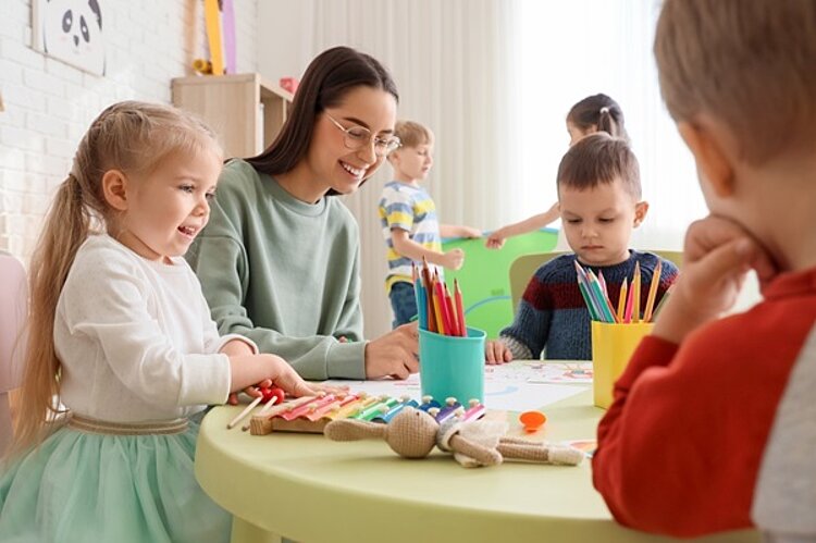 Kleine Kinder mit Erzieherin zeichnen gut gelaunt am Tisch im Kindergarten.