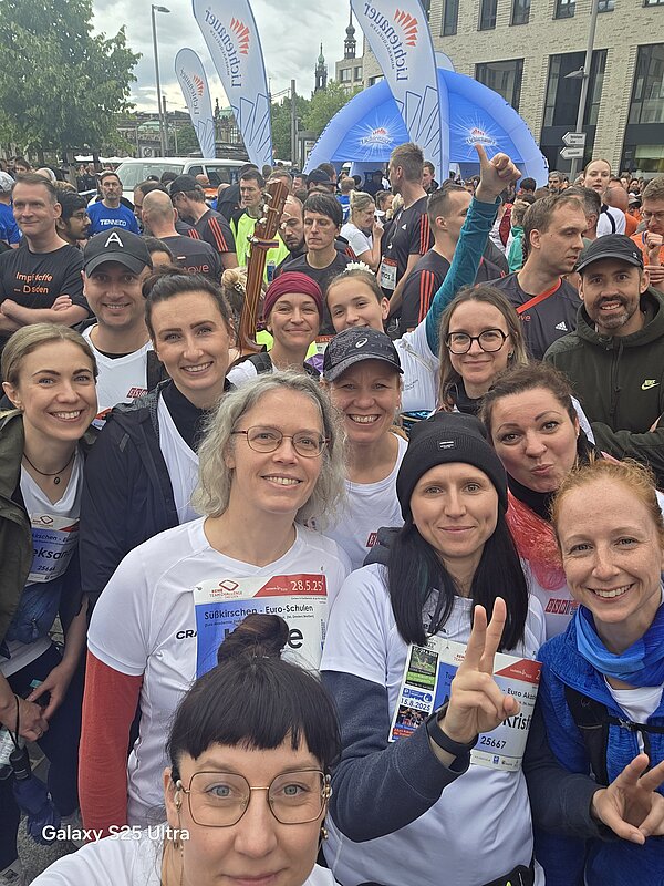 Selfie aus der Menschenmenge kurz vor dem Start. Die Teilnehmenden lachen in die Kamera, im Hintergrund sieht man blaue Zelte der Veranstaltung und viele weitere Läufer.