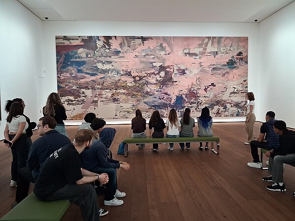 Die Auszubildenden sitzen und stehen in einem Ausstellungsraum mit hellem Holzfußboden und weißen Wänden. Im Hintergrund hängt ein großformatiges, abstraktes Gemälde mit überwiegend pastellfarbenen und erdigen Tönen. Einige Personen betrachten das Kunstwerk, während andere miteinander sprechen oder zuhören. Die Atmosphäre wirkt ruhig und konzentriert.