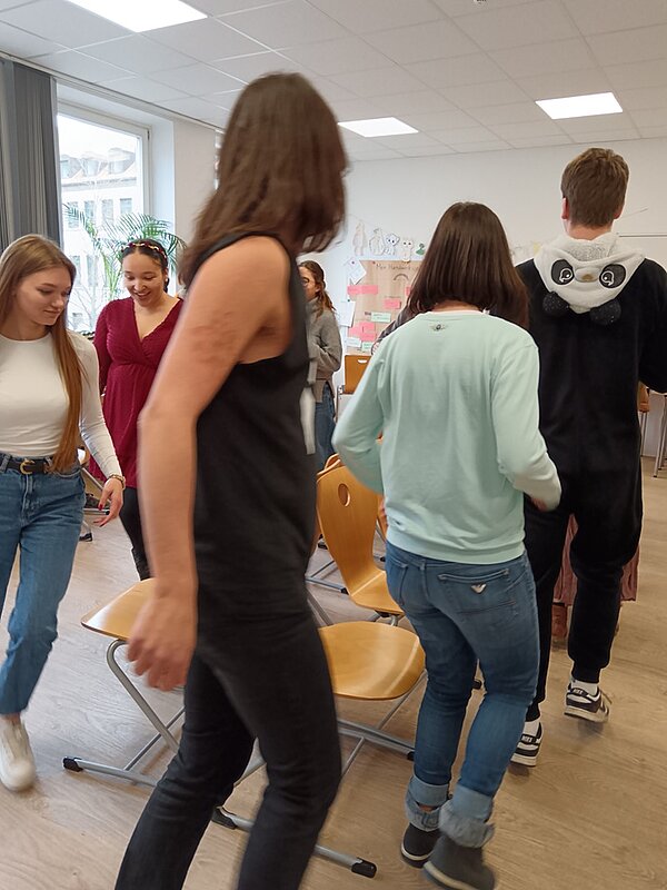 Studierende spielen "Die Reise nach Jerusalem" und gehen im Kreis an den aufgestellten Stühlen entlang.