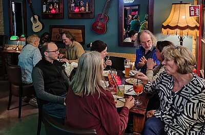 Mehrere Personen sitzen dicht beisammen an einem Tisch im Pub, essen und trinken Bier oder bunte Cocktails und unterhalten sich lebhaft, im Hintergrund hängen Gitarren und leuchten Tischlampen.